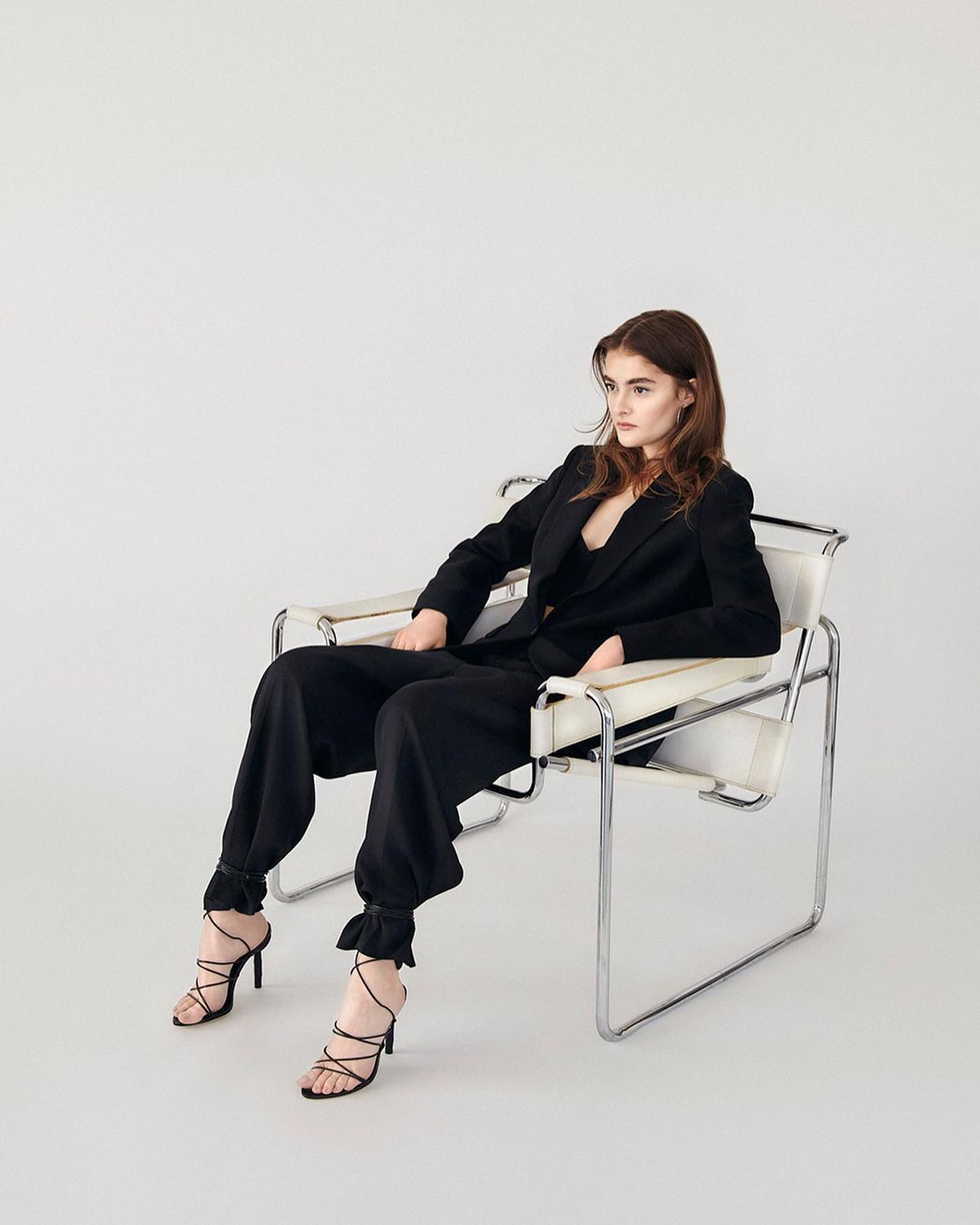 A woman with long brown hair sits in a white chair, wearing a black blazer, black pants, and strappy black high heels in a studio.