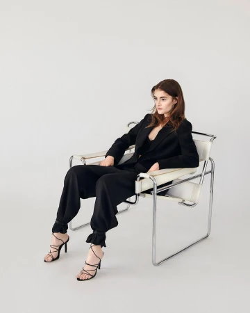 Model seated in a chrome and white leather armchair wearing a black pantsuit and strappy black high-heeled sandals against a white wall.