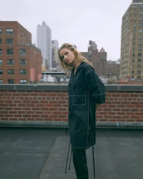 Model wearing dark coat with white topstitching standing on a New York rooftop against city skyline