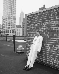 Woman in white suit leaning against brick wall on New York rooftop with skyline