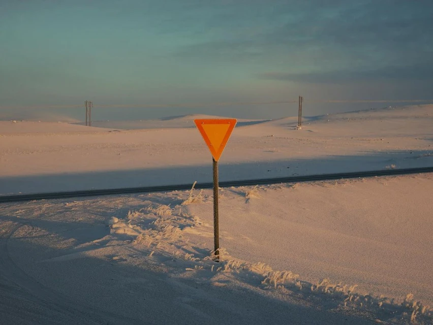 Yield sign along a paved road in a snowy, open landscape under a blue sky with power lines in the background at dusk or dawn.