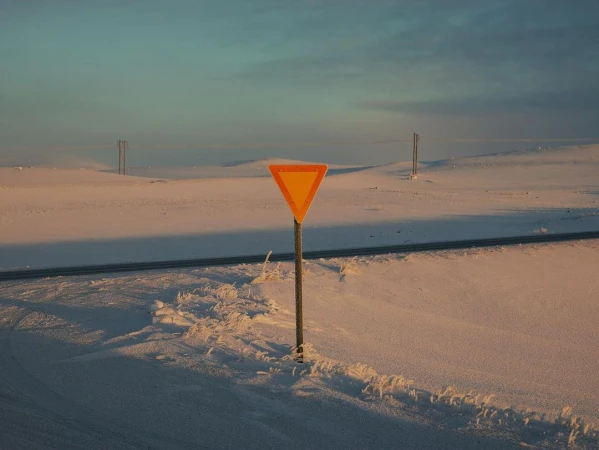 Yield sign along a paved road in a snowy, open landscape under a blue sky with power lines in the background at dusk or dawn.