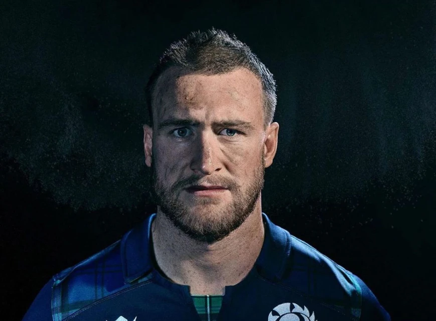 Close-up portrait of an adult male with dirty skin, wearing a blue and green plaid rugby shirt against a dark background.