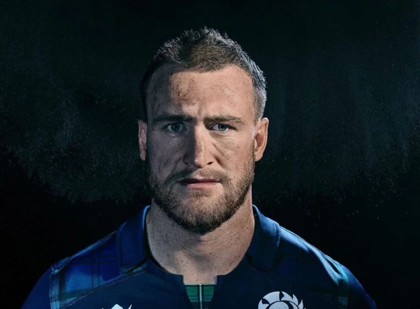 Close-up portrait of an adult male with dirty skin, wearing a blue and green plaid rugby shirt against a dark background.