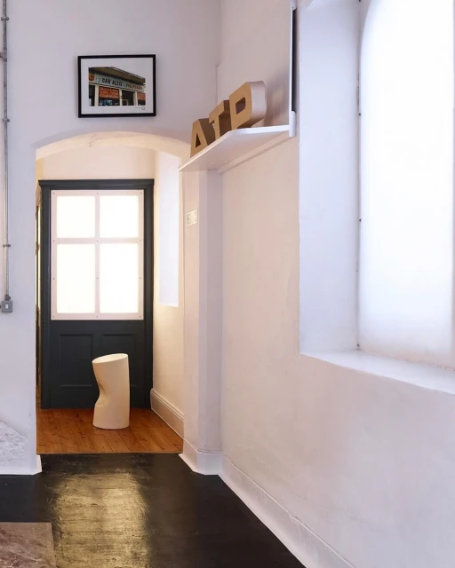 Minimalist interior hallway with white walls, black floor, natural wood stool, shelf with letters, and framed building photograph.