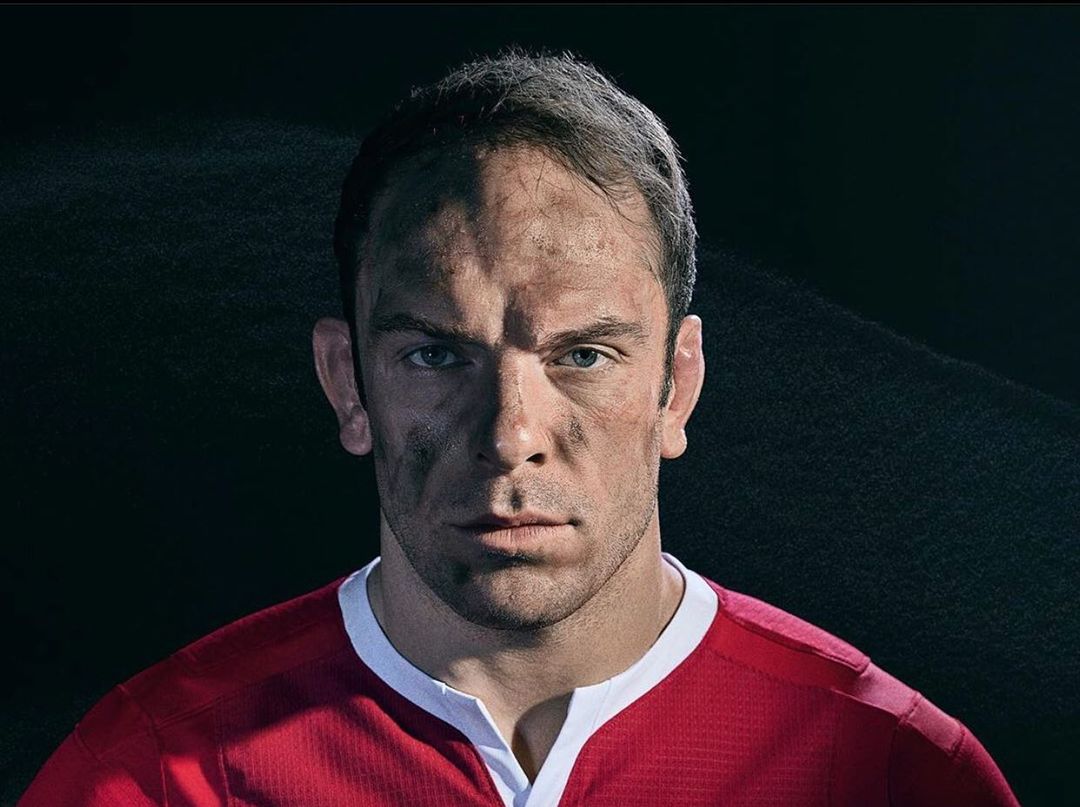 Close-up portrait of a man in a red athletic shirt with a determined expression, showing signs of dirt and exertion, set against a dark background.