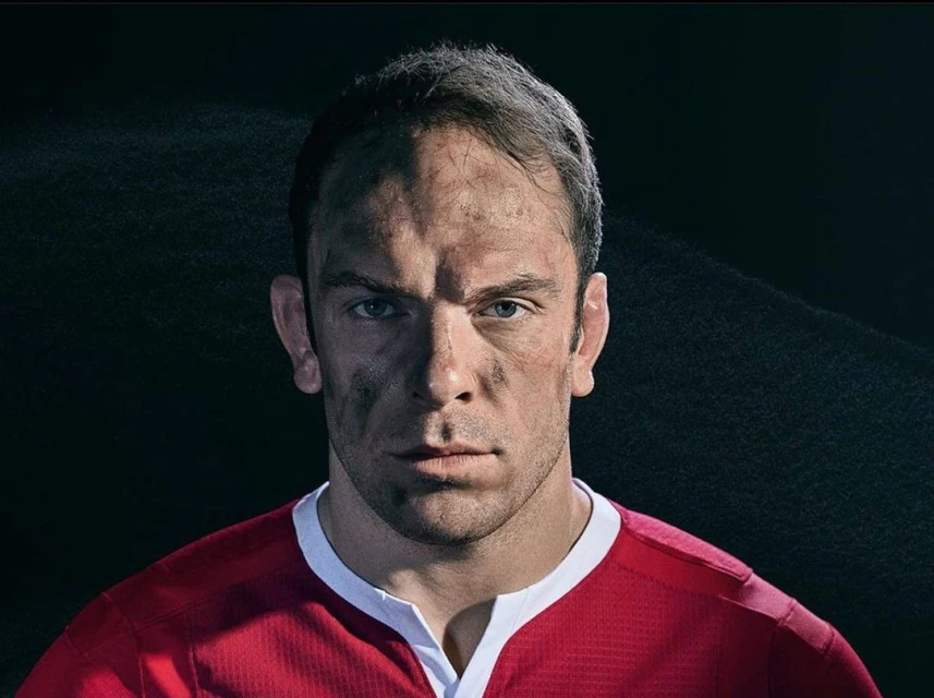 Close-up portrait of a man in a red athletic shirt with a determined expression, showing signs of dirt and exertion, set against a dark background.