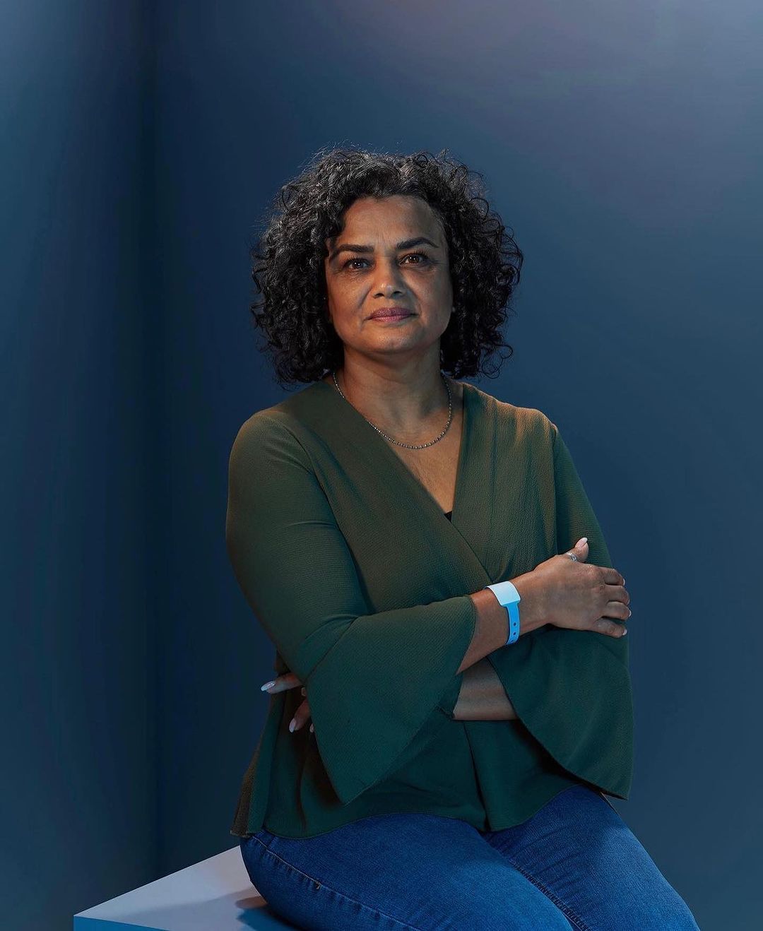 Portrait of a middle-aged woman with curly hair wearing a green top and jeans, seated indoors against a deep blue background.