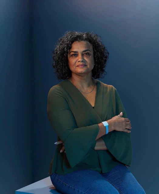 Portrait of a middle-aged woman with curly hair wearing a green top and jeans, seated indoors against a deep blue background.