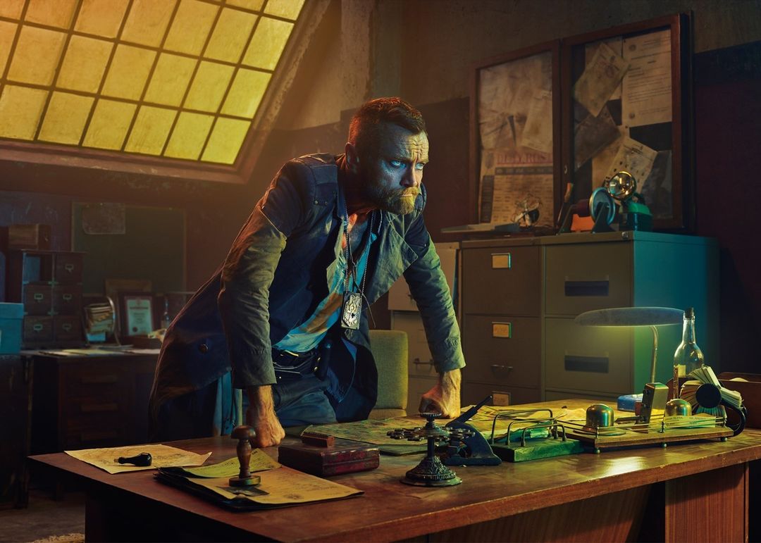 A man in a blue jacket leaning on a wooden desk in a dim office with papers and a desk lamp, looking serious.