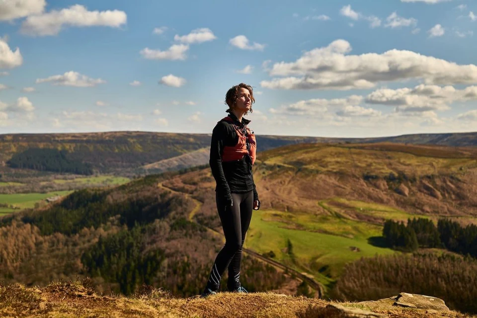 Female athlete stands on a hillside, wearing sports clothing, with a rural landscape under a partly cloudy blue sky in the background.