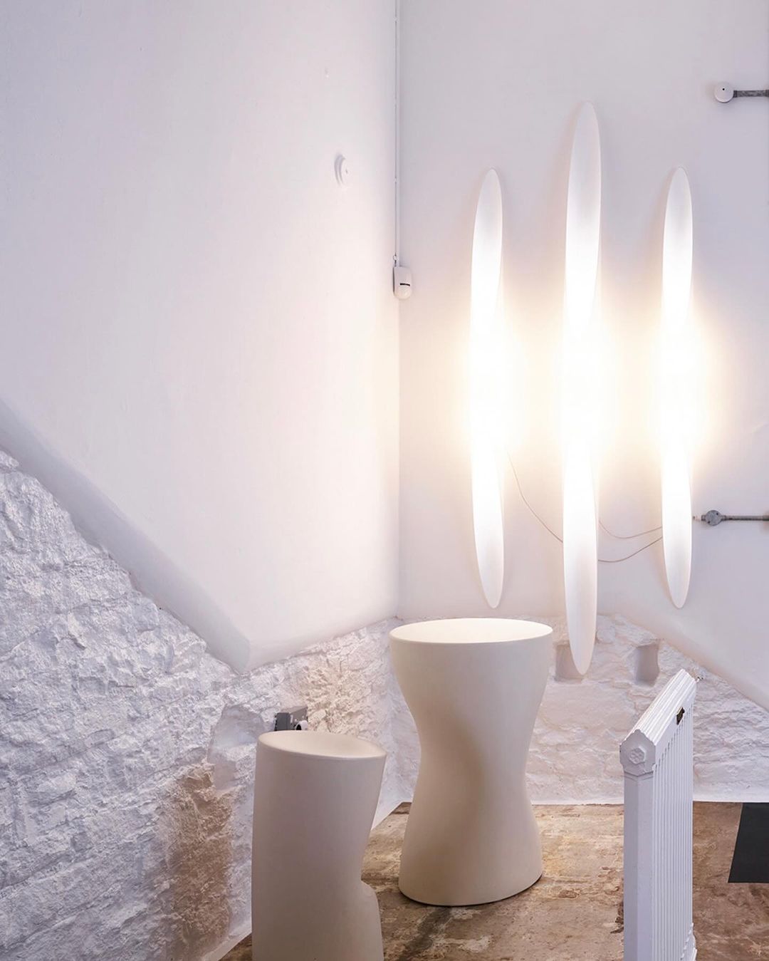 Minimalist interior space featuring two sculptural stools, radiant vertical light fixtures, and textured white walls, creating a clean and contemporary aesthetic.