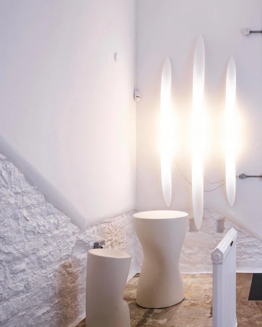 Minimalist interior space featuring two sculptural stools, radiant vertical light fixtures, and textured white walls, creating a clean and contemporary aesthetic.
