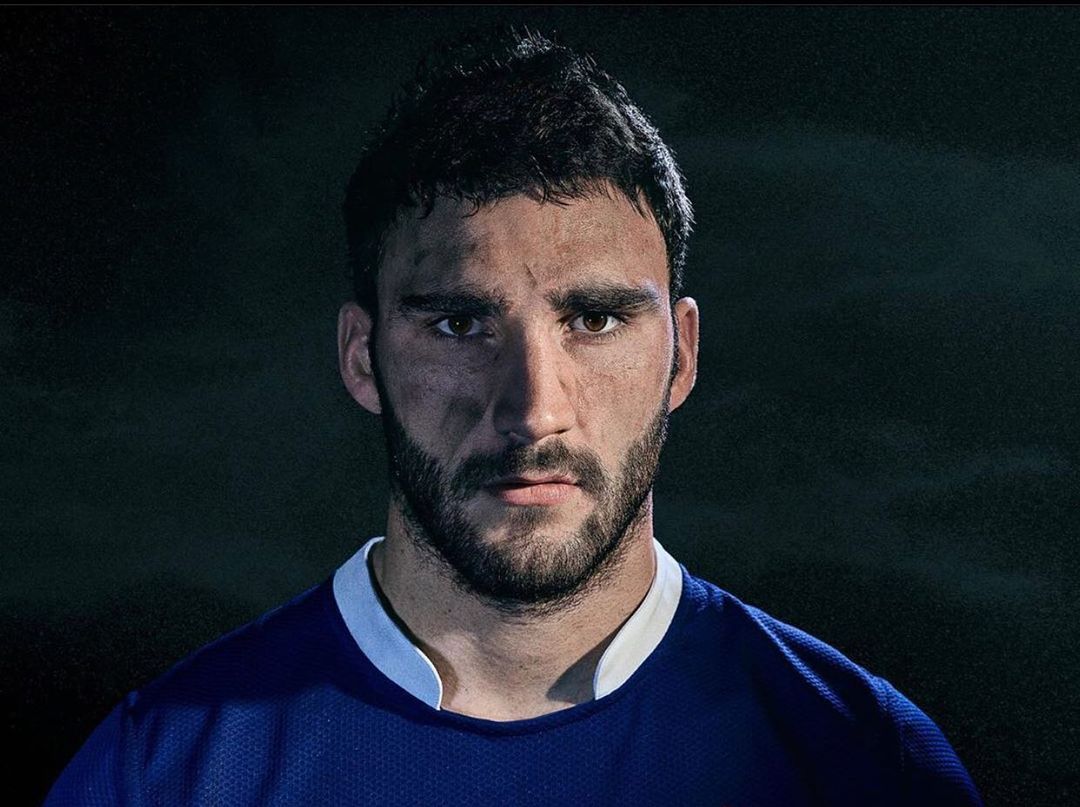 Headshot of a male athlete with dark hair and a beard, looking directly at the camera with dirt on his face and wearing a blue jersey.
