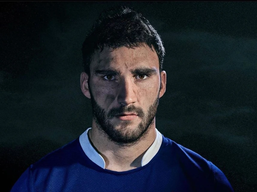 Headshot of a male athlete with dark hair and a beard, looking directly at the camera with dirt on his face and wearing a blue jersey.