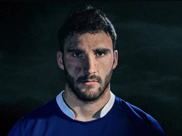 Headshot of a male athlete with dark hair and a beard, looking directly at the camera with dirt on his face and wearing a blue jersey.