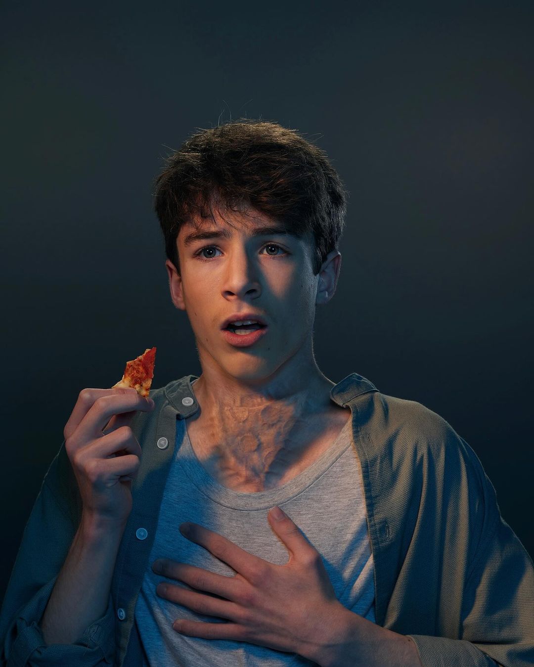 Young man in grey shirt and button-down looks surprised while holding a slice of pizza in a dimly lit indoor setting.