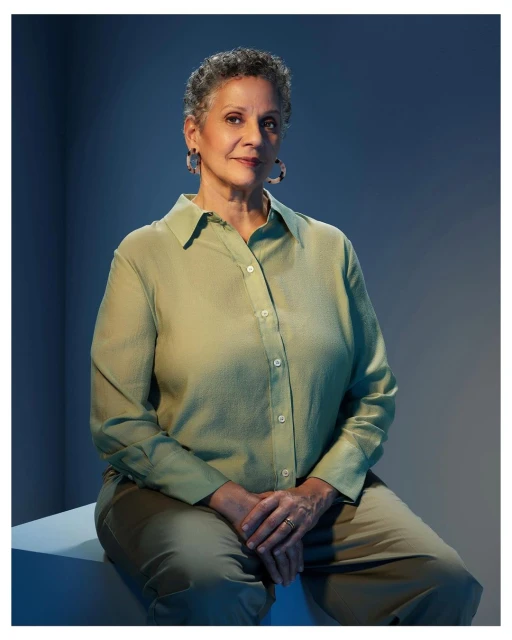 Mature woman in green shirt and khaki pants sits in a photo studio with blue background, tortoise earrings and hands clasped.