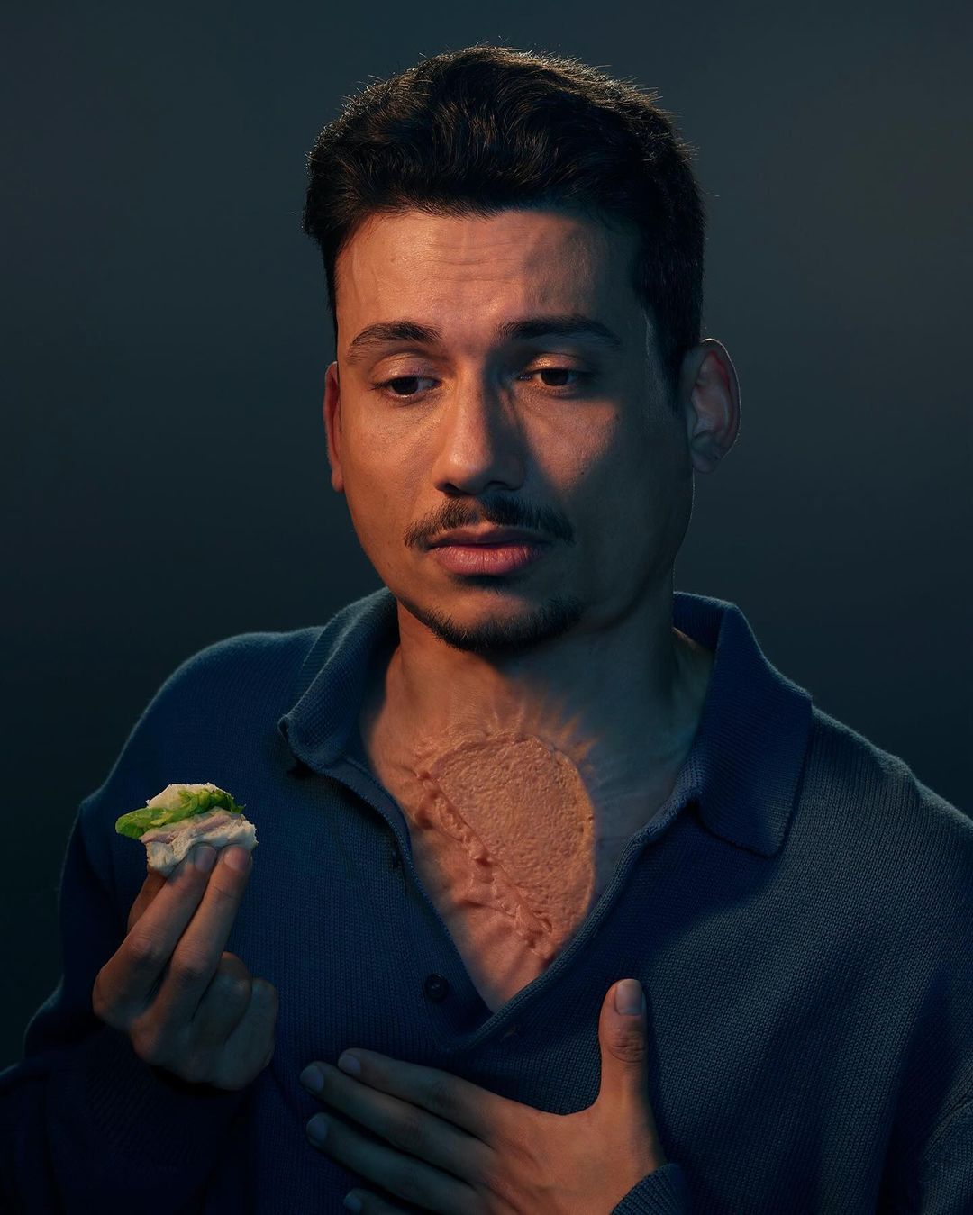 Portrait of a man with a scar on his chest, holding a sandwich, with a dark background and soft lighting creating a contemplative mood.