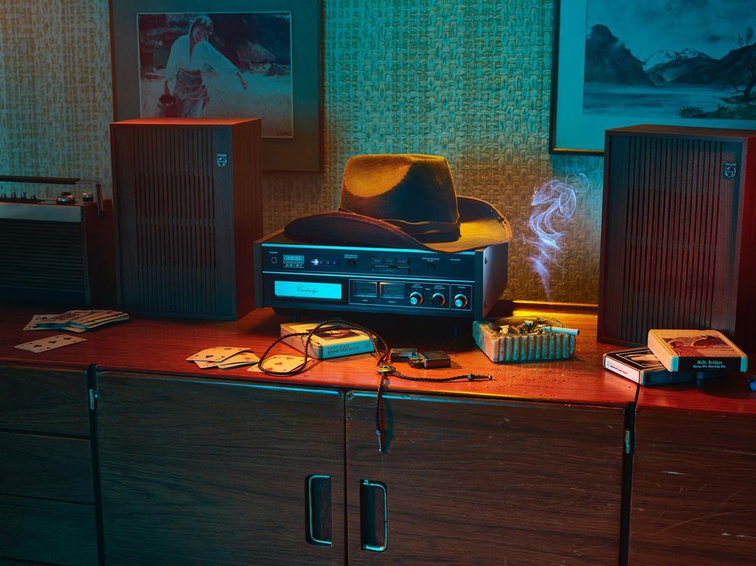 Retro audio system and accessories on a wooden dresser, featuring speakers, cassette player, cowboy hat, ashtray, playing cards, and art on wall.