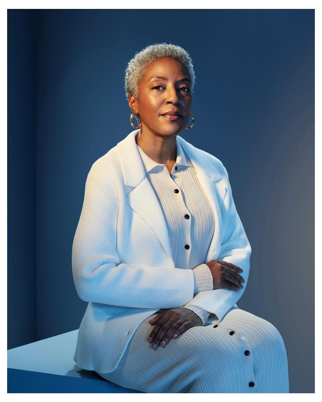 Portrait of a mature African-American woman with short gray hair in a cream-colored textured outfit, sitting indoors with a blue backdrop.
