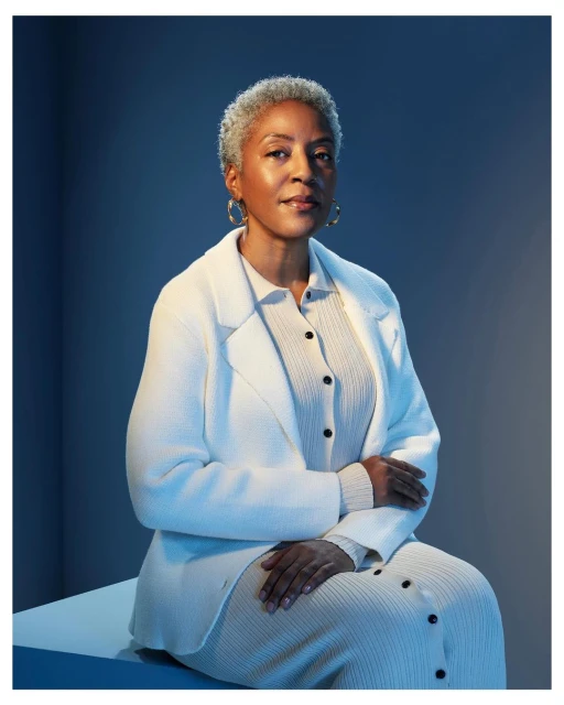 Portrait of a mature African-American woman with short gray hair in a cream-colored textured outfit, sitting indoors with a blue backdrop.
