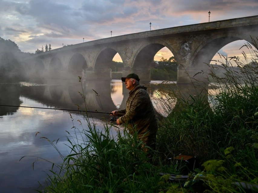 An adult male wearing a camouflage jacket and cap fishes with a fishing rod along a foggy river with a stone bridge in the background.