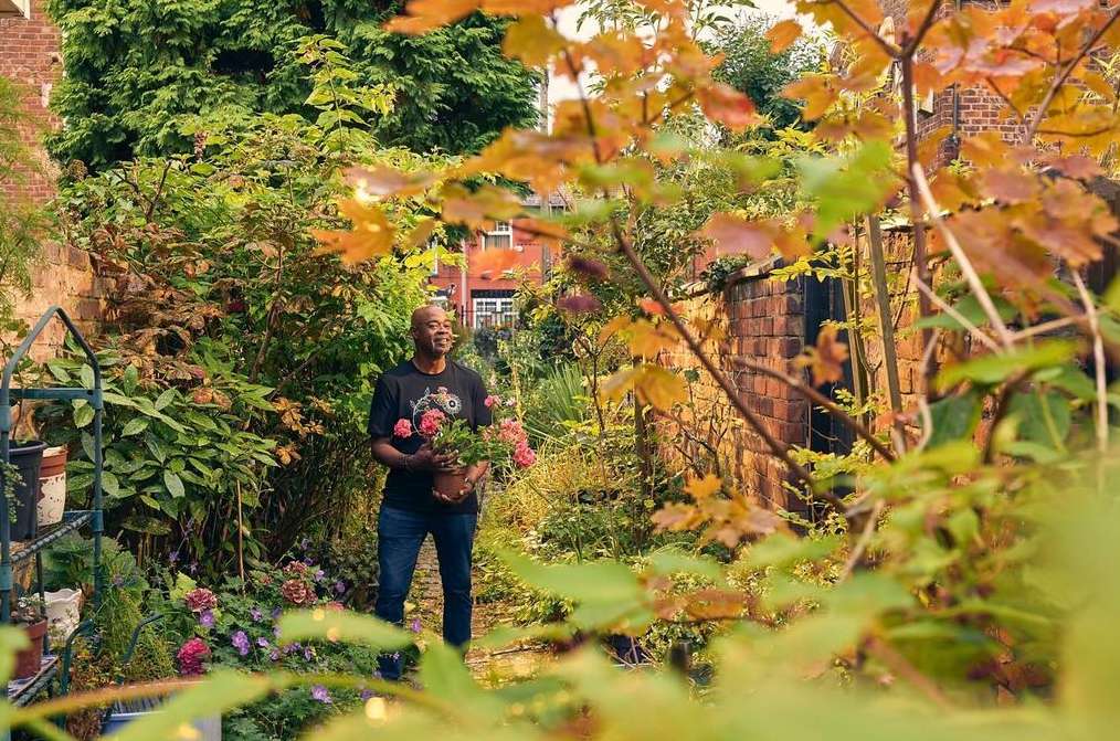 An adult male stands in a backyard garden, holding a potted plant with pink roses. The garden is lush, with a brick wall and flowers.