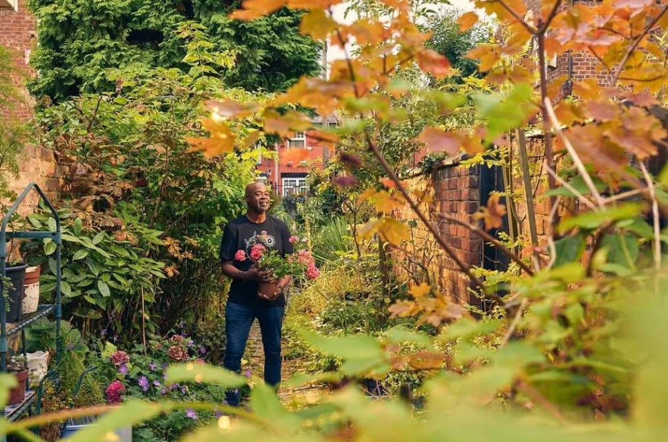 An adult male stands in a backyard garden, holding a potted plant with pink roses. The garden is lush, with a brick wall and flowers.