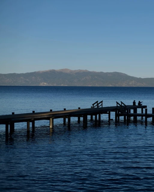 Scenic view of a wooden pier stretching into a calm, deep blue lake, with distant mountains under a clear sky; two people on the pier.