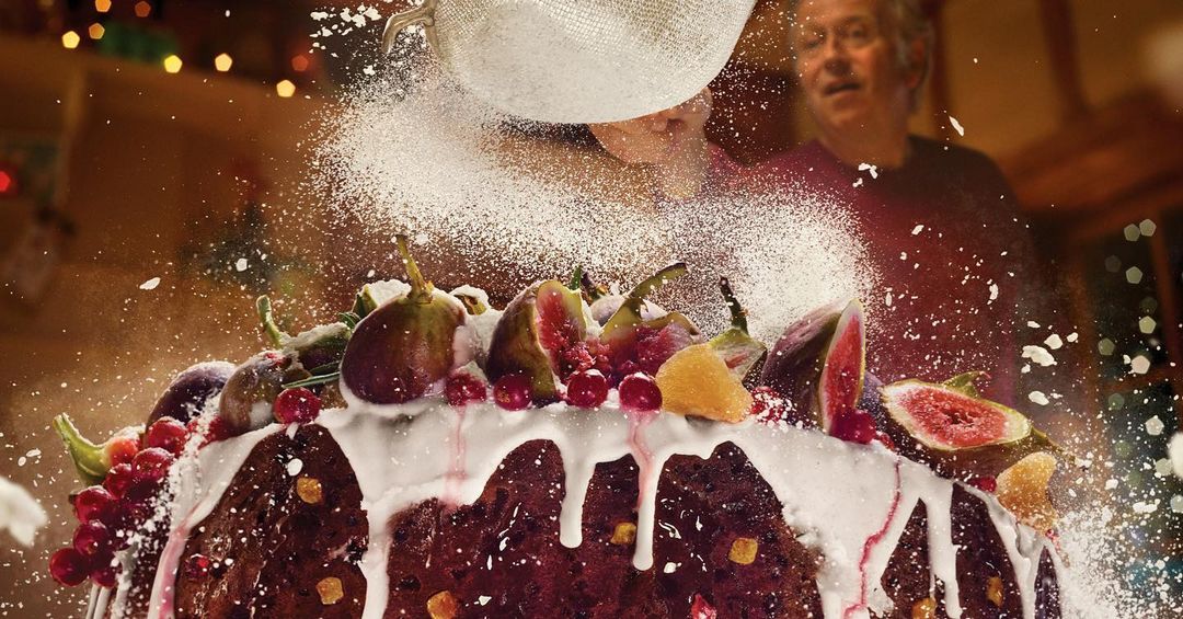 Iced fruitcake with figs and cranberries, dusted with powdered sugar being sifted from a metal sieve, with a blurred adult male in background.
