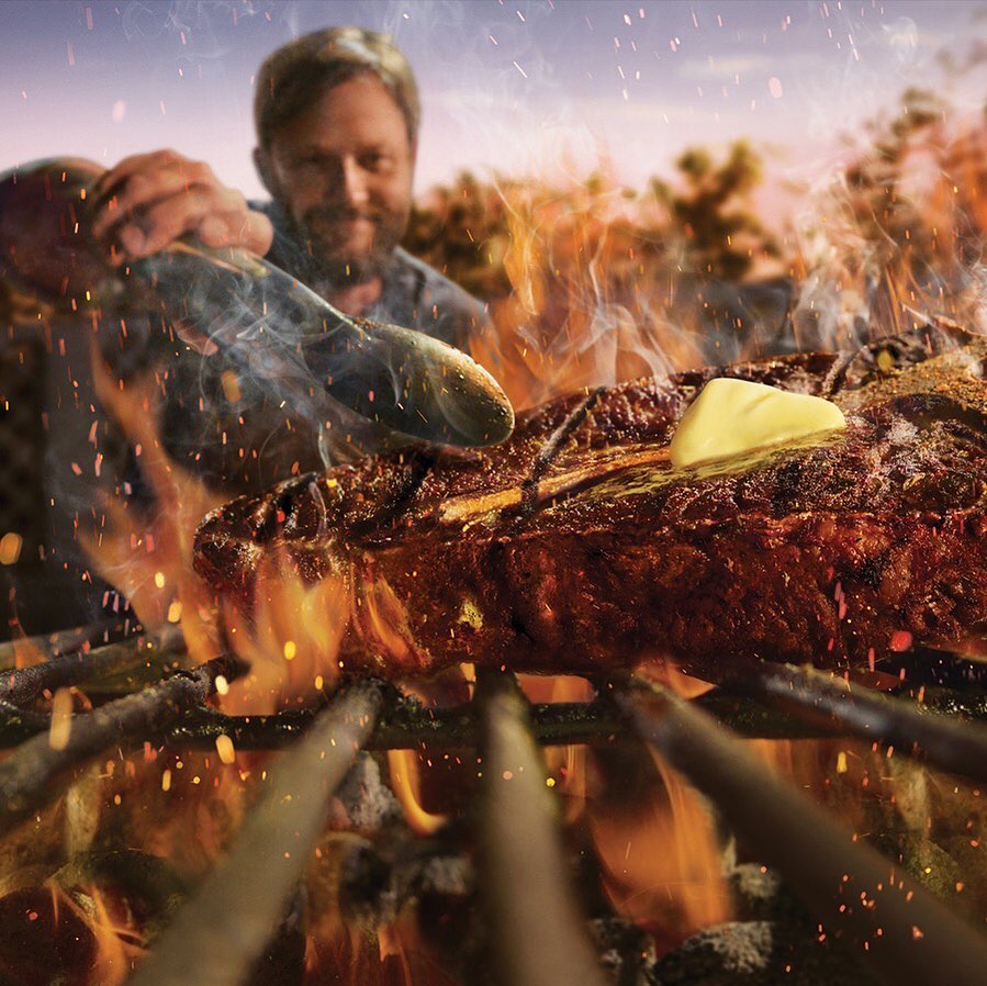 Close up of grilled steak with butter on skewers cooking over a fire, outdoors with a man visible in the background.