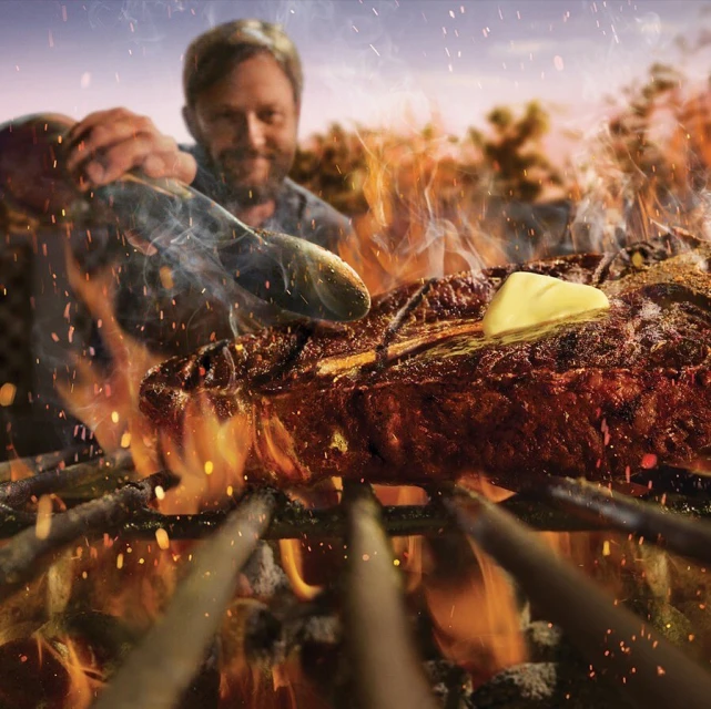 Close up of grilled steak with butter on skewers cooking over a fire, outdoors with a man visible in the background.