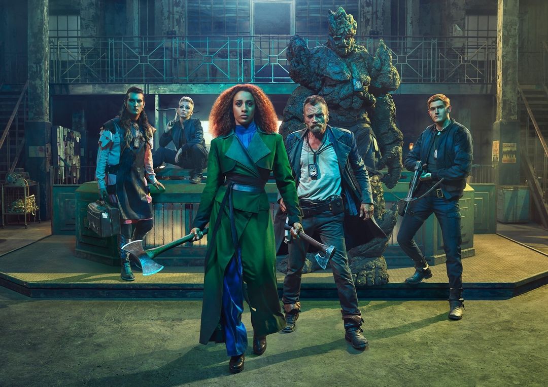 Group of five individuals stand with axes in an industrial-style location, featuring a large rock-like figure in the background.