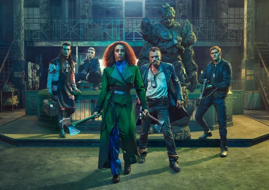 Group of five individuals stand with axes in an industrial-style location, featuring a large rock-like figure in the background.