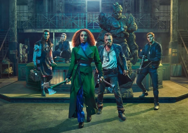 Group of five individuals stand with axes in an industrial-style location, featuring a large rock-like figure in the background.