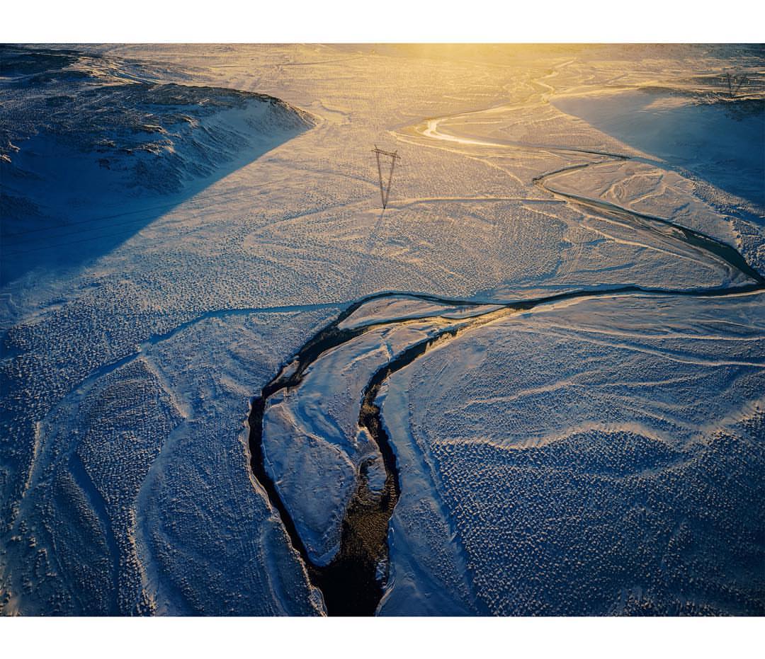 Aerial view of snowy landscape with winding river and overhead power lines under bright sun, capturing the stark beauty of nature.