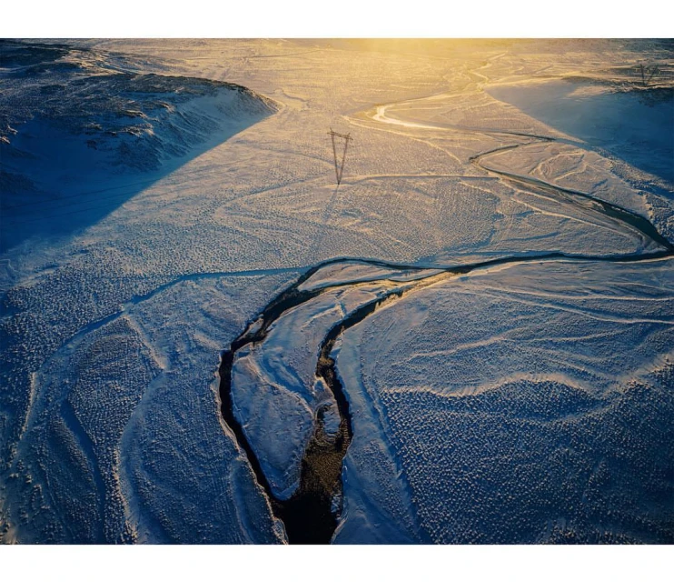Aerial view of snowy landscape with winding river and overhead power lines under bright sun, capturing the stark beauty of nature.