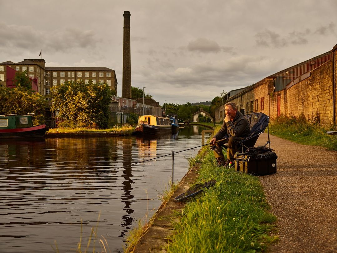 Man fishing in a canal with brick buildings and smokestack in the background during golden hour.
