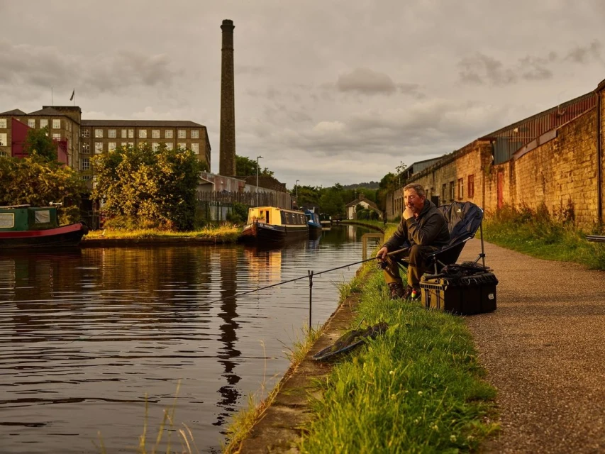 Man fishing in a canal with brick buildings and smokestack in the background during golden hour.