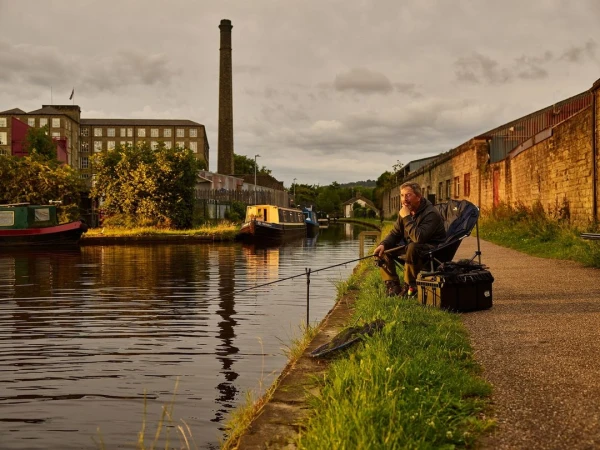 Man fishing in a canal with brick buildings and smokestack in the background during golden hour.
