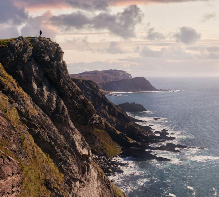 Person stands atop rocky cliff overlooking ocean with distant mountains and cloudy sky in scenic outdoor landscape shot.