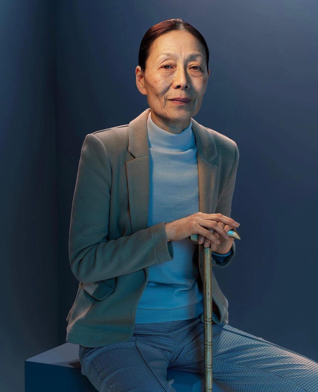 Elderly Asian woman in blazer and slacks seated indoors holding cane, gazing at camera in portrait framed by blue backdrop.