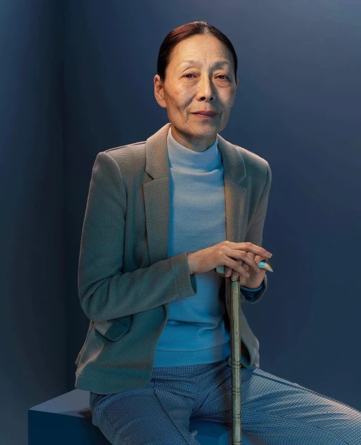 Elderly Asian woman in blazer and slacks seated indoors holding cane, gazing at camera in portrait framed by blue backdrop.