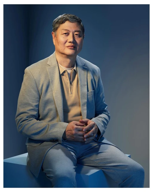 Portrait of an Asian adult male in a gray suit sitting with hands clasped, captured in a medium shot against a blue backdrop.