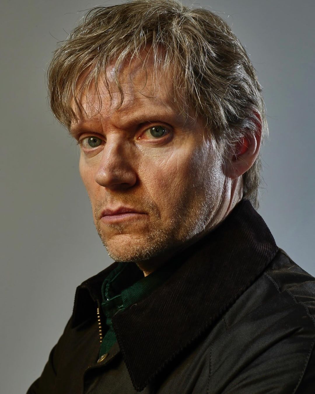 Portrait of a mature adult male with blond hair and a serious expression, wearing a dark jacket against a neutral grey background.