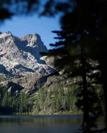 Scenic view of a rugged mountain range reflected in a calm lake surrounded by green trees, under a clear blue sky, framed by trees.