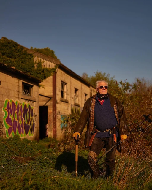 Elderly man stands outdoors in front of a dilapidated building, holding a shovel and wearing headphones and sunglasses in golden light.