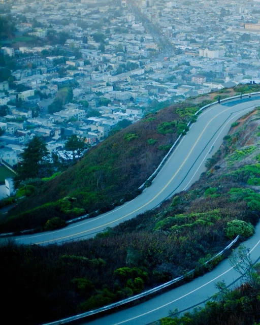 Aerial view of a winding road on a green hill leading towards a cityscape with many buildings under an overcast sky, creating a scenic vista.