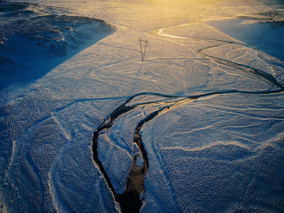 Aerial view of snowy landscape with mountains, a frozen river, and a transmission tower under bright sunlight, creating long shadows.